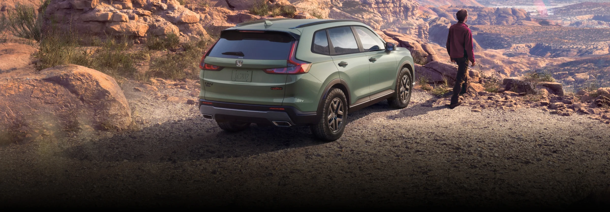 A green Honda CR-V parked on a rocky canyon overlook as a hiker takes in the view.