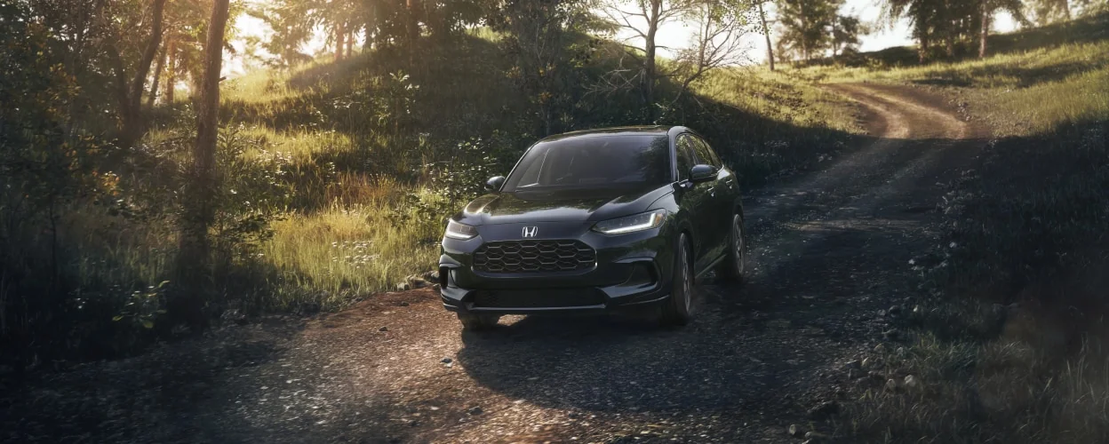 A dark HR-V driving down a gravel road through a sunlit forest.