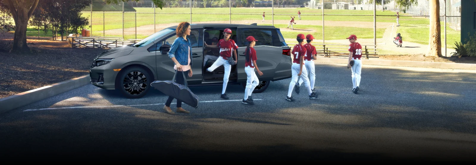 A youth baseball team climbing out of a grey Odyssey next to a ball diamond.
